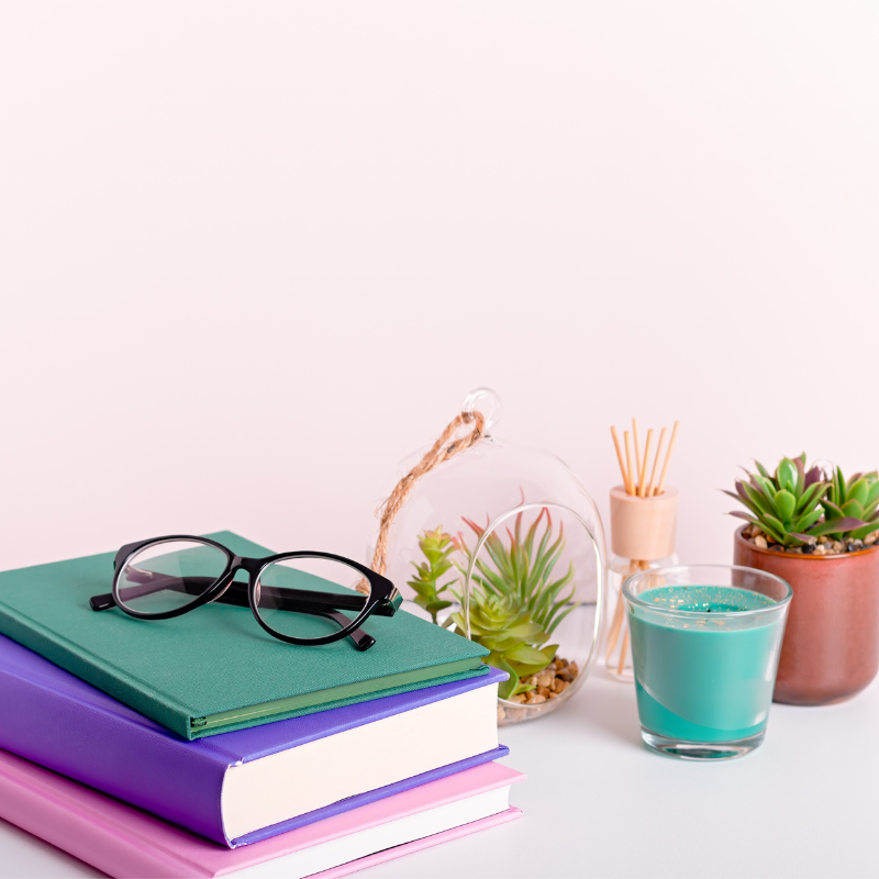 multi-coloured books stacked on a table with a pair of reading glasses and some pot plants